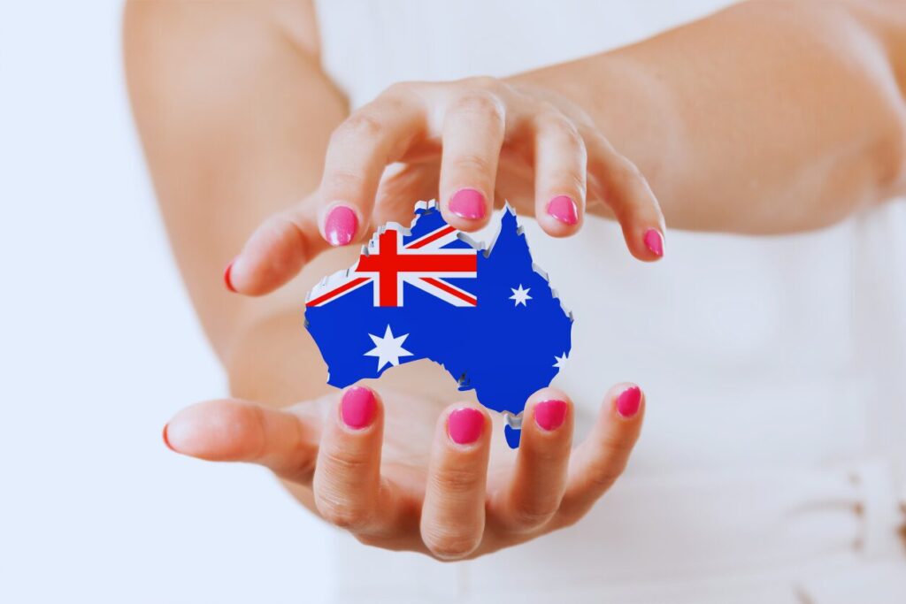A close-up of a person's hands with pink nail polish holding a 3D cutout map of Australia. The map is overlaid with the Australian national flag.